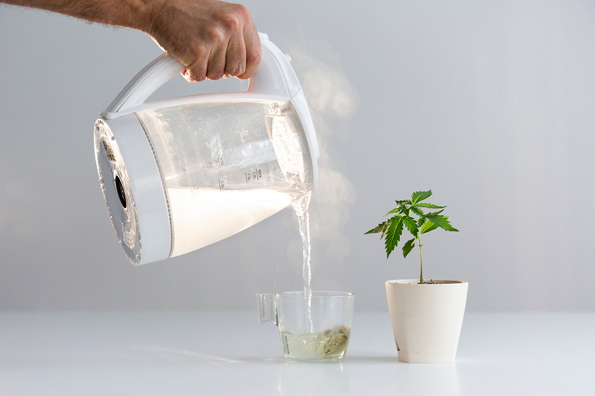 A clear kettle of boiled water being poured into a glass mug with a teabag in it.